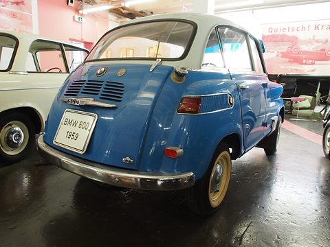 Rear three-quarter of a blue car with white roof on a dark museum floor, chrome bumper and red tail lamps. Opens a larger view
