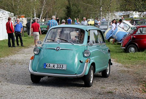 Turquoise bubble-shaped car seen head-on on a gravel track, driver behind the windscreen, visitors and Isettas behind. Opens a larger view