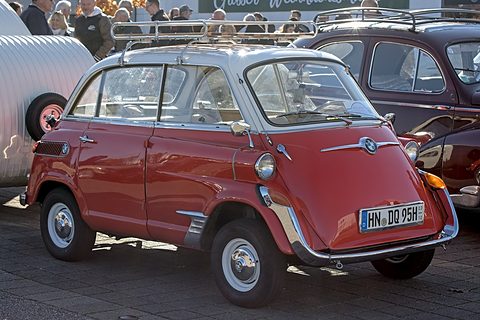 Red small car with white roof and chrome roof rack on paving, a crowd and a dark saloon behind. Opens a larger view