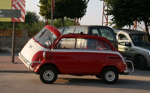 Small red car with white nose panel raised like a hatch, parked at a kerb beside a van and an estate car. Opens a larger view