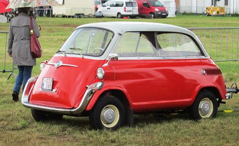 Red small car seen from its right side on grass, rain drops on the windows, vans and a fence behind. Opens a larger view