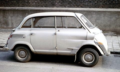 Pale grey bubble-shaped car parked at a kerb in front of a concrete wall topped with dark brick. Opens a larger view