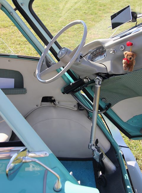 Close view inside a turquoise car: grey thin-rimmed wheel on a hinged column, speedometer, a teddy-bear mascot. Opens a larger view