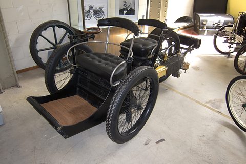 Black three-wheeled veteran car with buttoned leather seats and a wooden front footboard. Opens a larger view