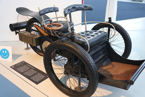 Black three-wheeled veteran car with tandem seats and an exhaust pipe on a white plinth with a text panel. Opens a larger view