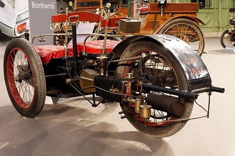 Rear of a three-wheeled veteran car showing a finned cylinder, exhaust pipe and large rear mudguard. Opens a larger view