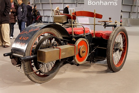 Side of a three-wheeled veteran car with a finned horizontal engine, belt pulley and red seats. Opens a larger view