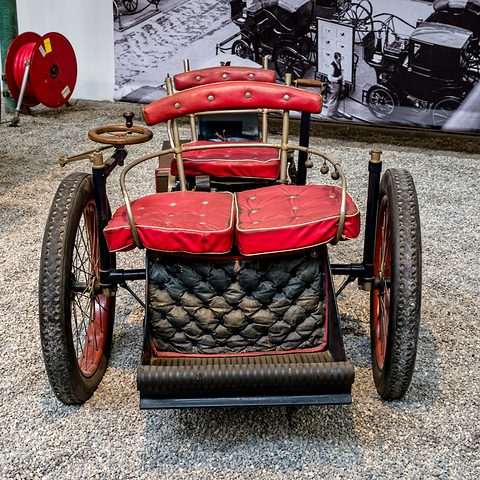 Head-on view of a three-wheeled veteran car with red seats and a quilted black front panel on gravel. Opens a larger view
