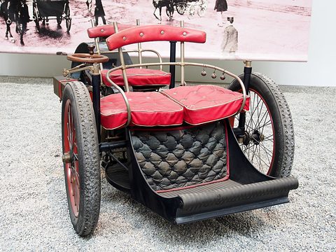 Front of a three-wheeled veteran car with quilted black leather front panel and red seats. Opens a larger view