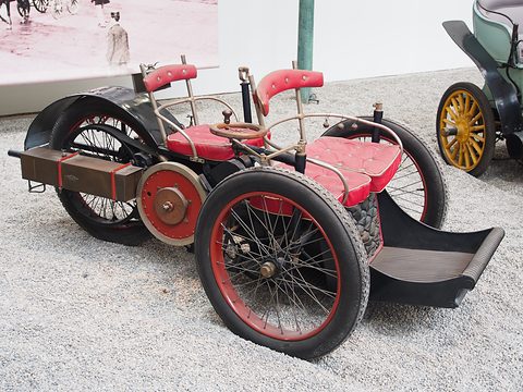 Three-wheeled veteran car with two red seats in tandem and a long front footboard on pale gravel. Opens a larger view