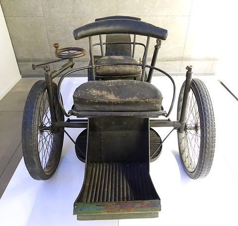 Front of a black three-wheeled car with a slatted front footboard and a leather seat on a white plinth. Opens a larger view