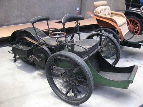 Black and green three-wheeled veteran car with two tandem seats and a front footboard on a pale floor.