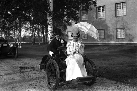A woman in a white dress with a parasol and a man in a top hat on a three-wheeled car. Opens a larger view