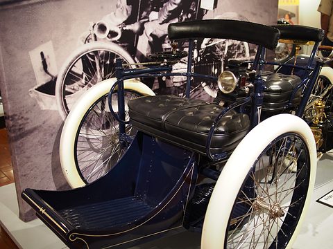 Close front view of a three-wheeled car with white tyres, a front footboard and buttoned seats. Opens a larger view