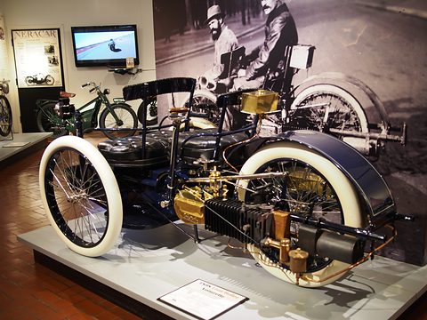 Side view of a dark blue three-wheeled car with white tyres and brass fittings before a large old photograph. Opens a larger view