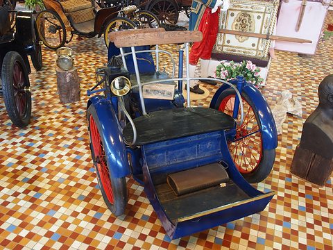 Front of a blue three-wheeled car with a wooden footboard and red wheels on a tiled floor. Opens a larger view