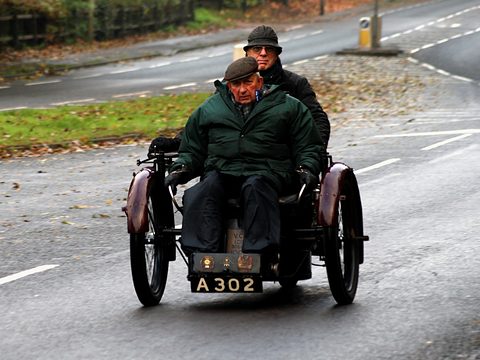 Two men in caps and coats aboard a small three-wheeled car on a wet road. Opens a larger view