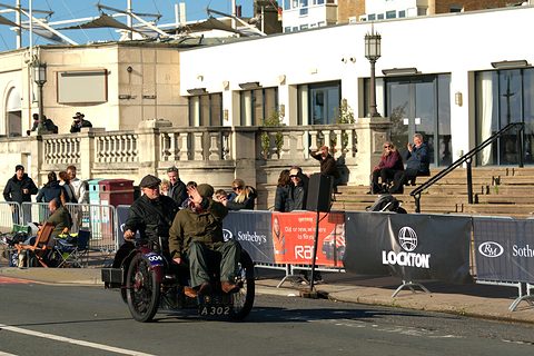 Small three-wheeled car with two men aboard passing sponsor barriers and spectators on a seafront. Opens a larger view
