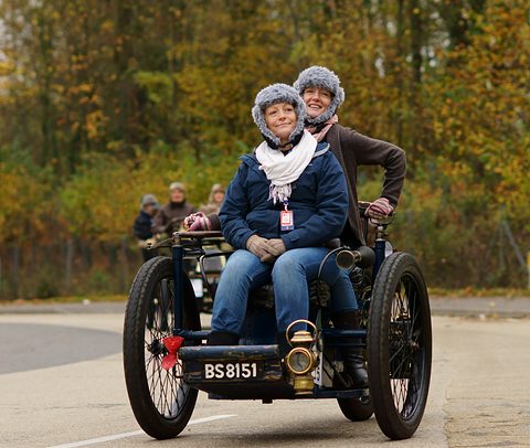 Two women in fur hats aboard a small black three-wheeled car, autumn trees behind. Opens a larger view