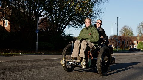 Two people aboard a small three-wheeled car on a suburban road in bright low sun. Opens a larger view