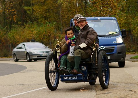 Three-wheeled veteran car with a child in front and a man behind, a van following on the road. Opens a larger view