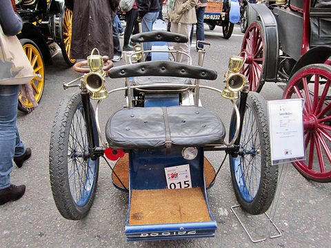 Front of a blue three-wheeled veteran car with brass lamps and a leather seat, visitors around it. Opens a larger view
