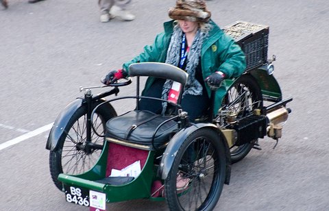 Looking down on a green three-wheeled car with a woman in a fur hat at the tiller and a basket behind. Opens a larger view