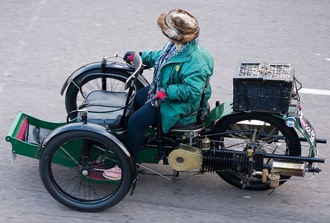 Side of a green three-wheeled car with a horizontal engine, a basket and a driver in a fur hat. Opens a larger view