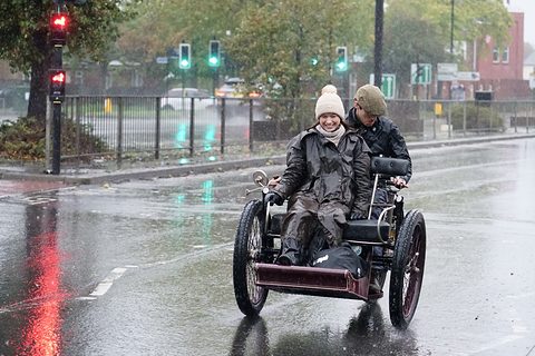 Two people in waterproofs aboard a small three-wheeled car on a flooded road, traffic lights behind.