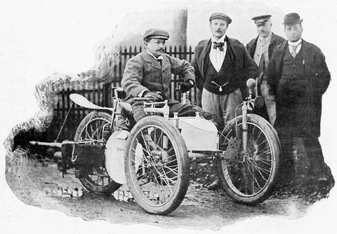 A man in a cap seated on a three-wheeled car, three men in hats and suits standing beside it. Opens a larger view