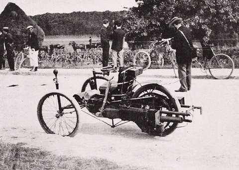 A small three-wheeled car on a sandy road, men and bicycles standing behind.