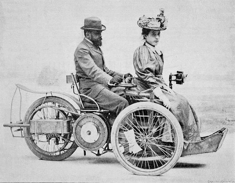 A bearded man in a bowler hat drives a three-wheeled car with a woman in a feathered hat in front.