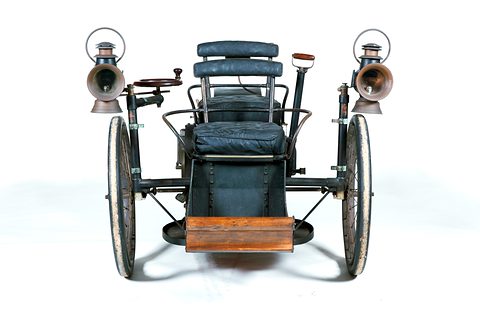 Head-on view of a three-wheeled veteran car with two carriage lamps and a wooden footboard on white. Opens a larger view
