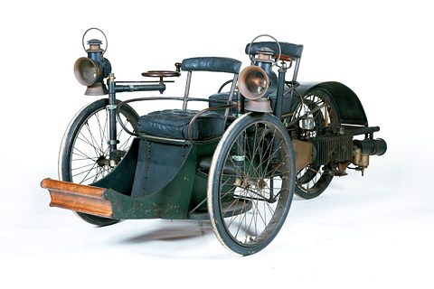 Dark green three-wheeled veteran car with carriage lamps and tandem leather seats against white. Opens a larger view