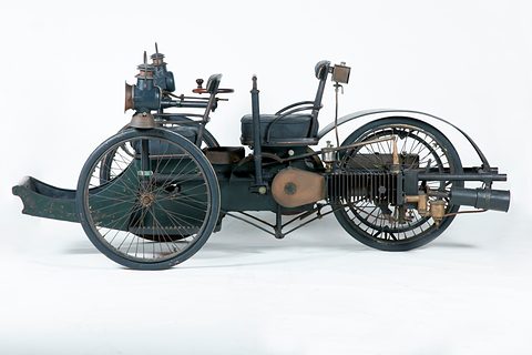 Side view of a three-wheeled car with a finned horizontal engine at the back against white. Opens a larger view