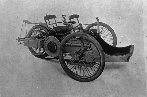 A three-wheeled veteran car with two tandem seats and a long front footboard, shown against a plain backdrop. Opens a larger view