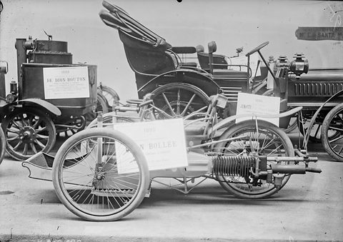A three-wheeled veteran car with a horizontal engine and a placard reading ‘Bollée’, other old cars behind. Opens a larger view