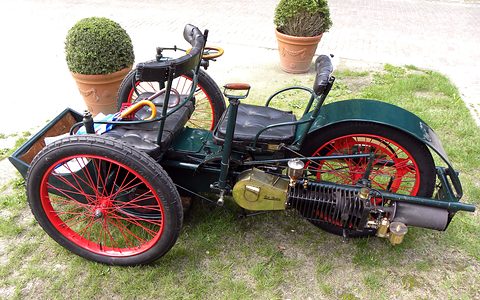 Dark green three-wheeled veteran car with red wheels and a horizontal engine on grass by potted box trees. Opens a larger view