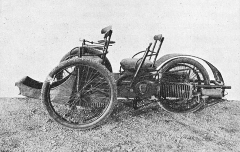 A three-wheeled veteran car with two seats in tandem standing on stony ground. Opens a larger view