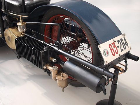 Close view of a finned engine cylinder, brass fittings and a rear mudguard with a race plate. Opens a larger view