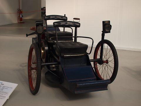 Front of a dark blue three-wheeled veteran car with tandem leather seats on a grey floor. Opens a larger view