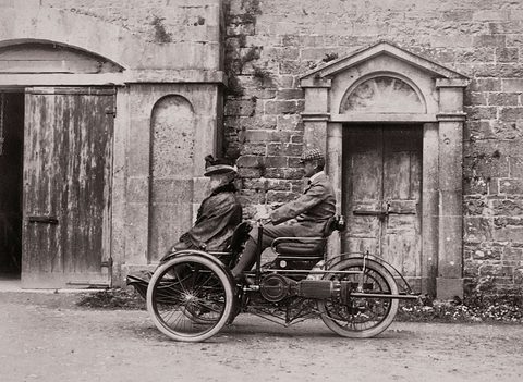 Two men in coats seated on a small three-wheeled car in a stone-walled stable yard. Opens a larger view