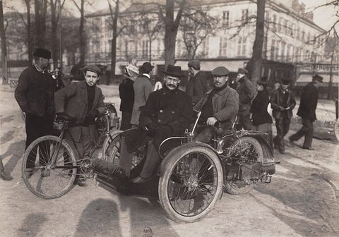 A man on a three-wheeled car with a passenger in front, onlookers in coats on a street behind. Opens a larger view