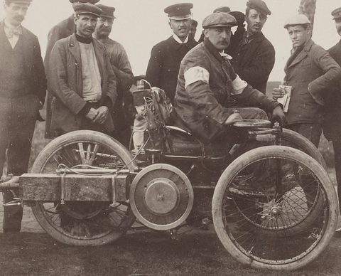 A man in a cap with an armband seated on a three-wheeled car, a crowd of men around him. Opens a larger view