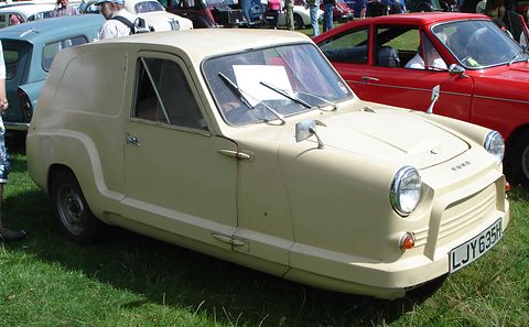 Cream three-wheeled van with blind rear sides parked on grass, a red car behind.