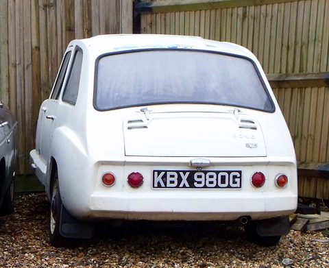 White small car from behind, four round tail lamps and louvres on the engine lid, on gravel.