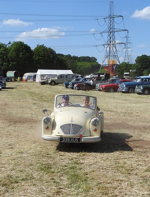 Cream three-wheeler driving towards the camera across dry grass under a pylon and blue sky. Opens a larger view