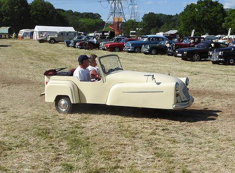 Cream open three-wheeler with two occupants crossing dry grass, a line of parked cars behind. Opens a larger view