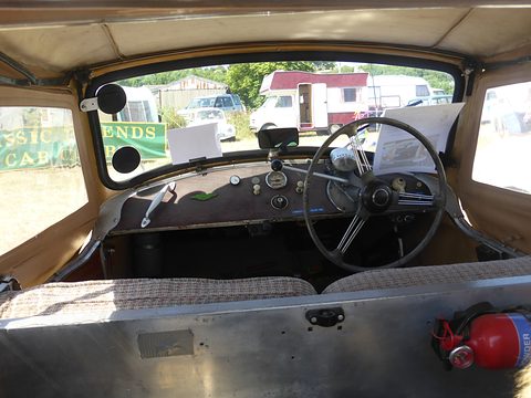 Looking forward under a beige hood to a worn painted dash, a steering wheel and a fire extinguisher. Opens a larger view