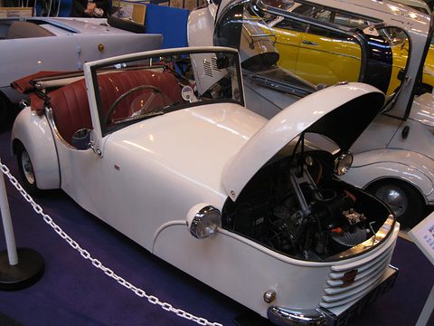 White open three-wheeler with its nose tilted forward behind a rope, a yellow car beyond. Opens a larger view